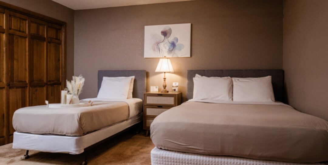 Two beautifully made beds with taupe duvets and white pillows with taupe walls and soft lighting
