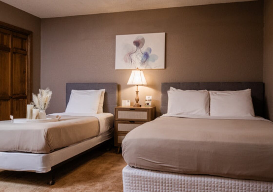 Two beautifully made beds with taupe duvets and white pillows with taupe walls and soft lighting