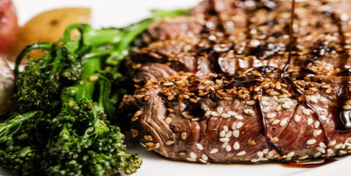 A sesame-crusted sirloin steak sits on a white plate with broccolini next to it