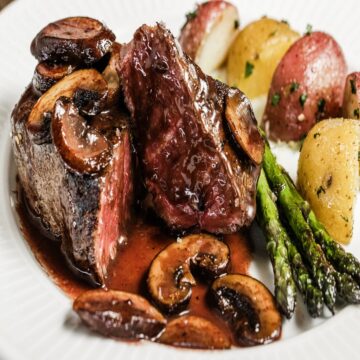 A juicy beef tenderloin steak sits on a white plate next to roasted asparagus and roasted potatoes