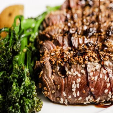 A sesame-crusted sirloin steak sits on a white plate with broccolini next to it