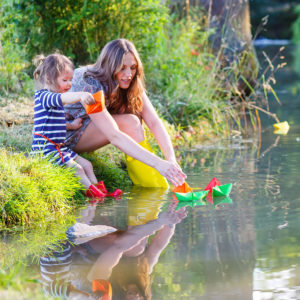 Young beautiful woman and her little daughter playing together with paper boats in a river. Creative leisure with kids.