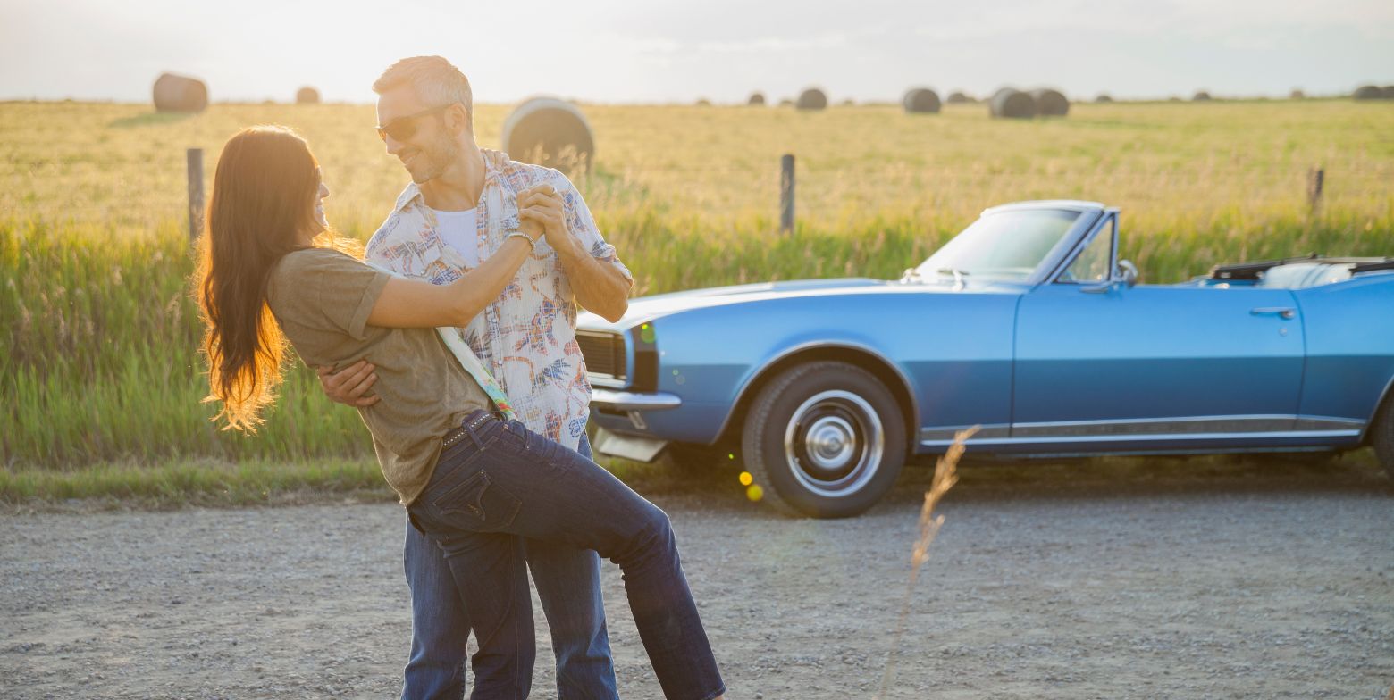 A couple dancing on a country road, their car parked on the side next to a field