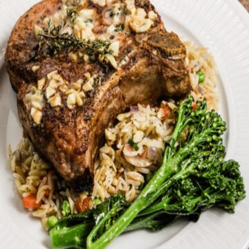 A thick grilled pork chop lays on a bed of rice with roasted broccolini next to it on a white plate