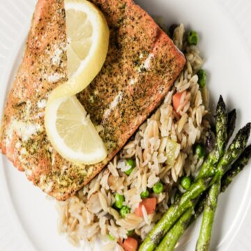 An herb crusted Salmon filet sits on a bed of rice with broccolini next to it on a white plate