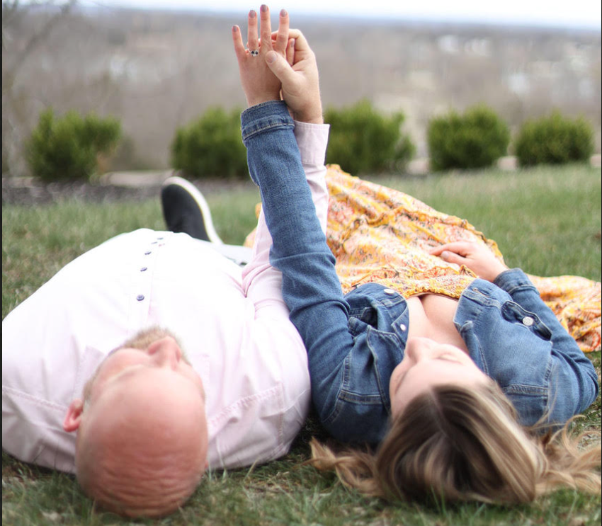 Engaged couple relaxing on the grass at Cedar Crest Lodge, lying side by side and admiring an engagement ring, with soft natural scenery in the background—capturing a joyful, intimate moment perfect for romantic getaways and proposal celebrations in Kansas.