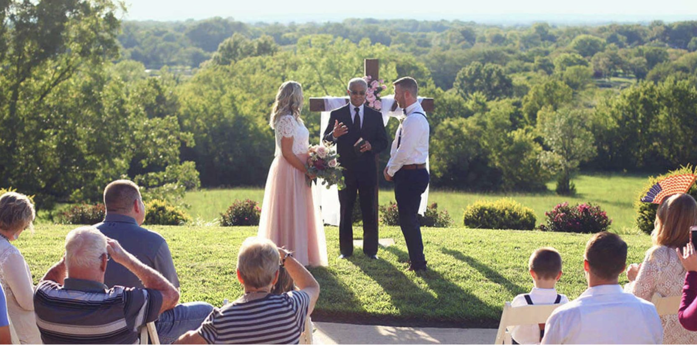 A bride in a lace top and blush pink skirt stands with a groom in a white shirt and navy trousers during an outdoor wedding ceremony at Cedar Crest Lodge. They are positioned before a wooden cross draped in white fabric and pink flowers, with a scenic backdrop of rolling green hills and lush trees under a bright, clear sky