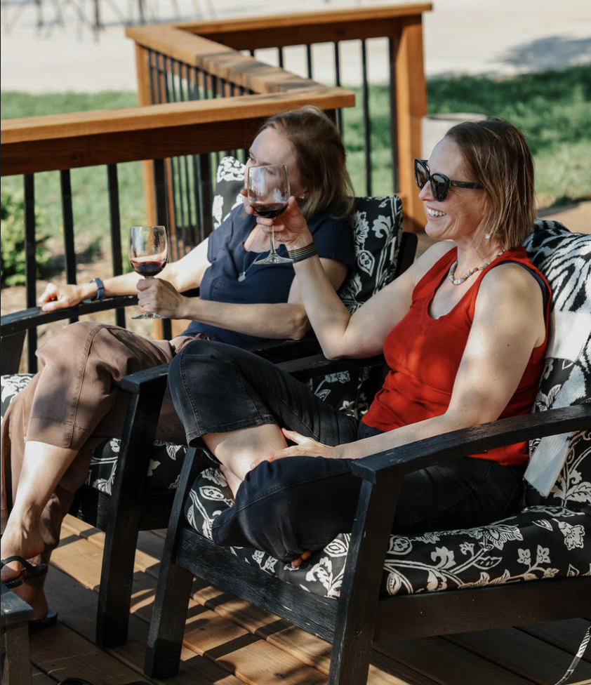 Two women smiling and relaxing on a sunny outdoor patio deck at Cedar Crest Lodge in Pleasanton, KS, comfortably seated in patterned armchairs while enjoying glasses of red wine during a girlfriend getaway trip.